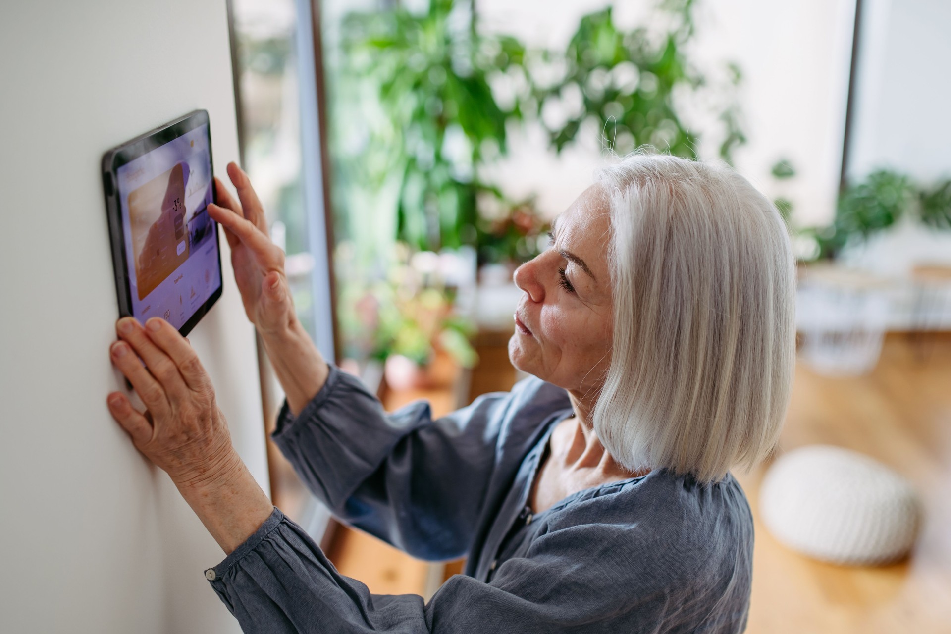 Mature woman adjusting smart thermostat, touch screen of smart home device. Older woman using smart technology at home.